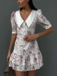 A woman wearing a vintage-style white floral dress with puff sleeves, a large collar, and ruffled hem, posing against a gray background.