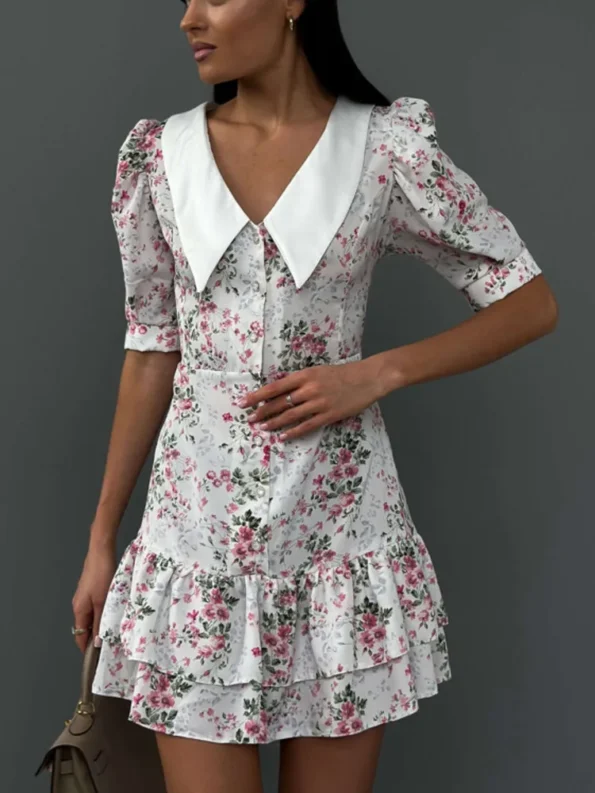 A woman wearing a vintage-style white floral dress with puff sleeves, a large collar, and ruffled hem, posing against a gray background.