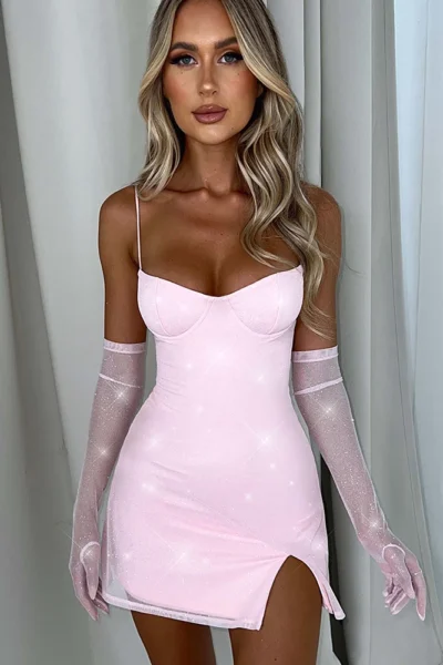 A woman wearing a glamorous light pink spaghetti strap mini dress with a thigh slit, paired with sheer matching gloves, posing elegantly indoors.